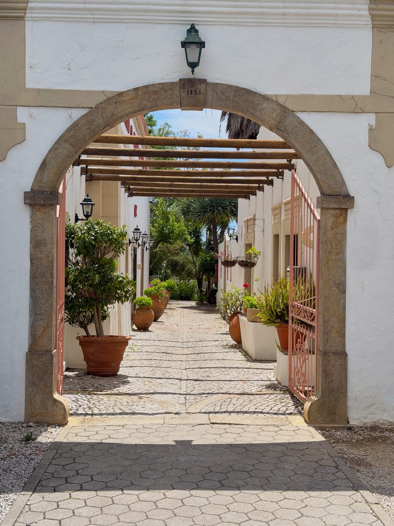 IMG_9128_web - Monte da Palhagueira Stone archway with date 1893 framing a sunny courtyard of white walls, wooden pergola beams, and potted plants.